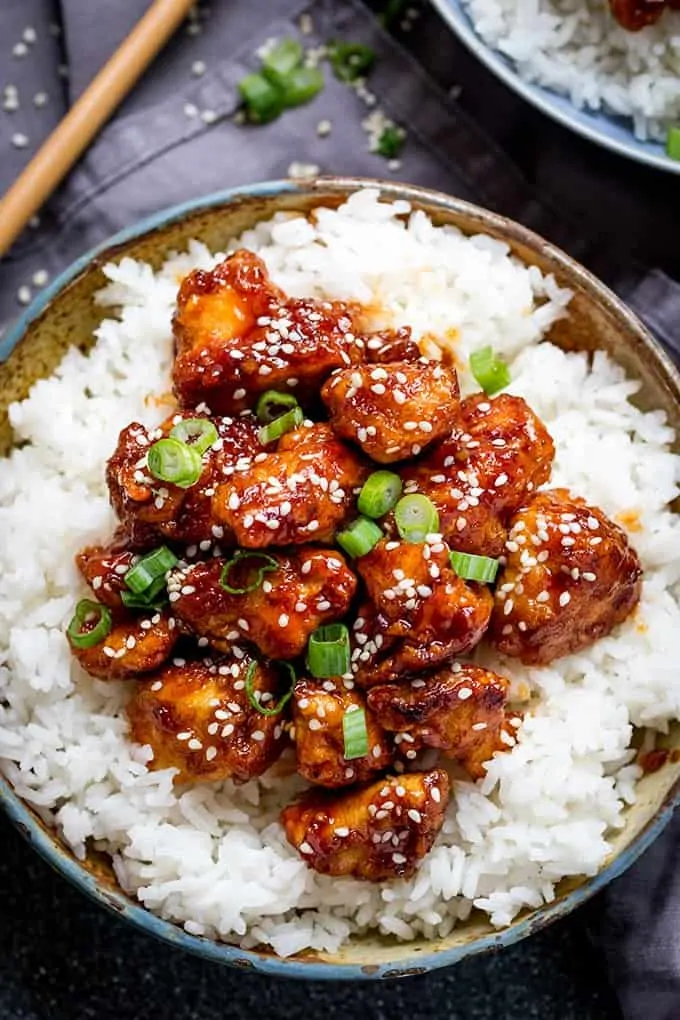 Overhead picture of Crispy Sesame Chicken with a Sticky Asian Sauce on a bed of rice in a stone bowl with a dark background with chopsticks in the top left corner