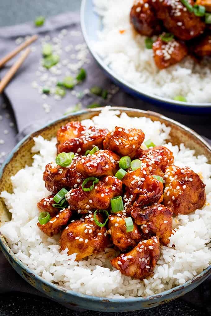 Portrait photo of Crispy Sesame Chicken with a Sticky Asian Sauce on a bed of rice in a stone bowl with another bowl and chopsticks in the background