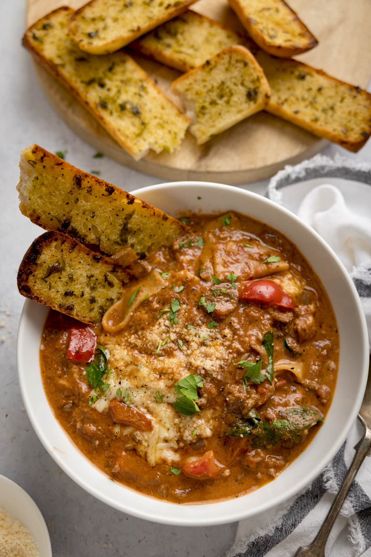 Overhead image of a white bowl filled with lasagne soup with pieces of garlic bread taken out. The bowl is on a light background next to a white napkin. There are more garlic bread sliced in the background.