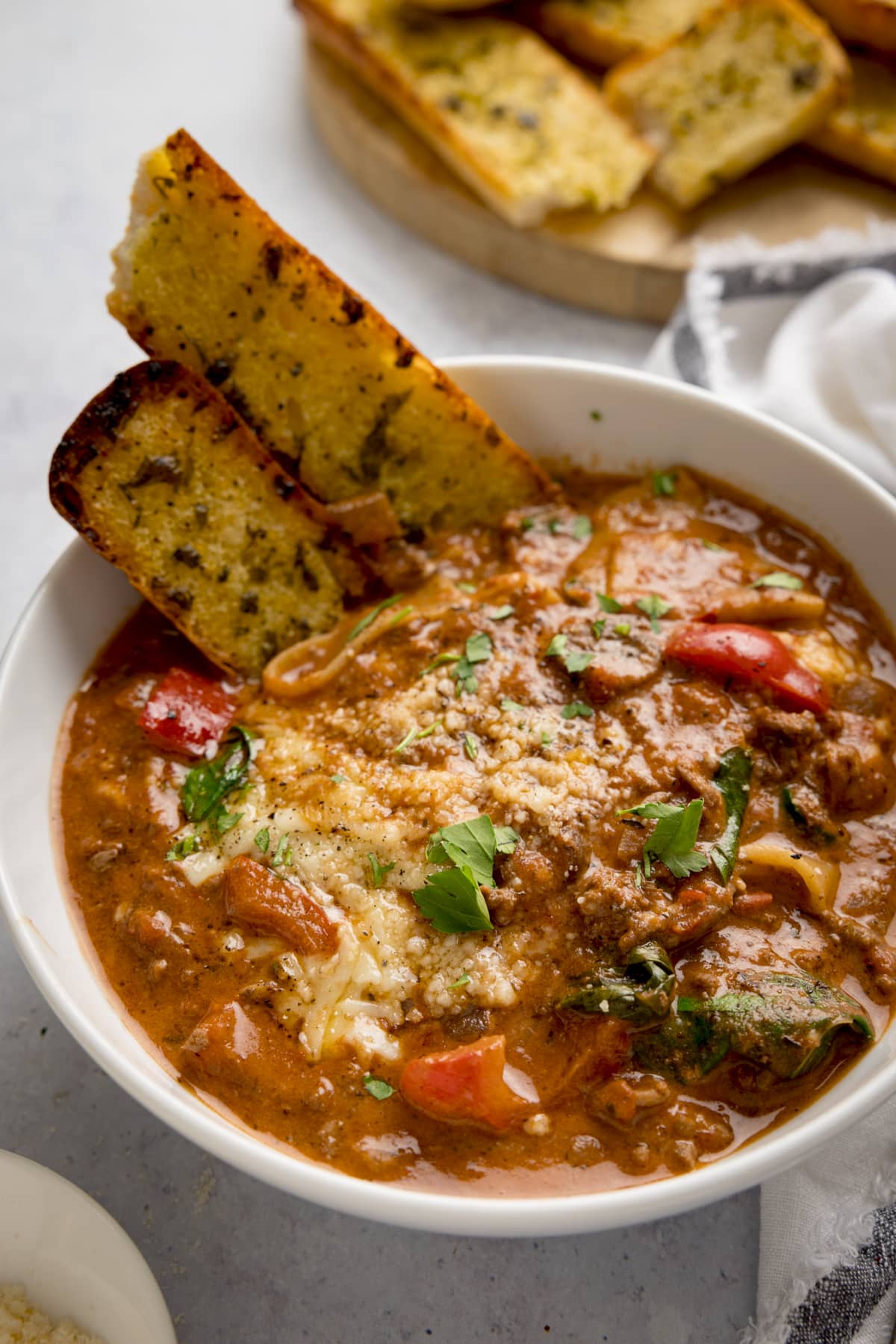 White bowl filled with lasagne soup with pieces of garlic bread taken out. The bowl is on a light background next to a white napkin. There are more garlic bread sliced in the background.