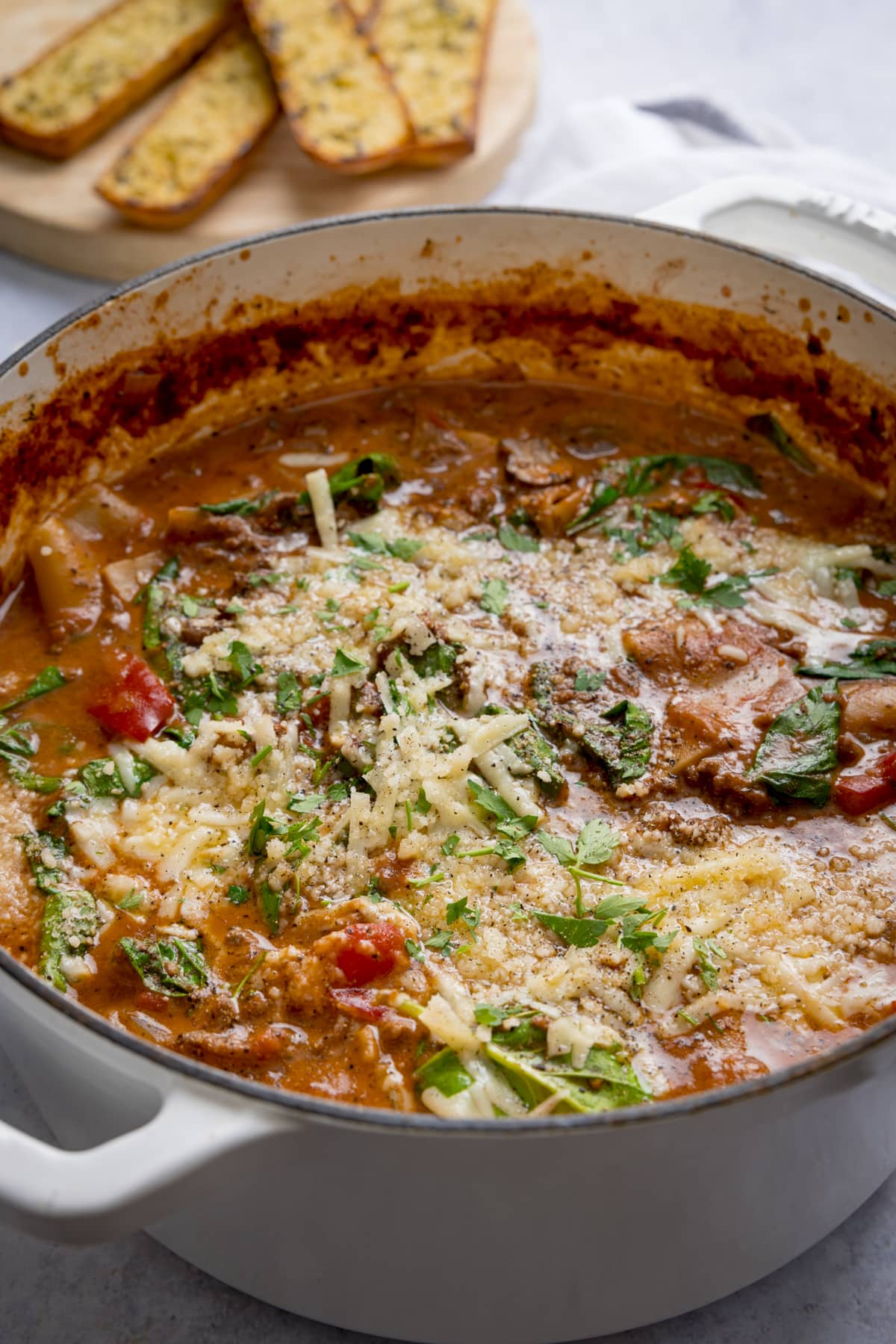 large white cast iron pan filled with lasagne soup on a light background. There are slices of garlic bread just in shot at the top of the frame.