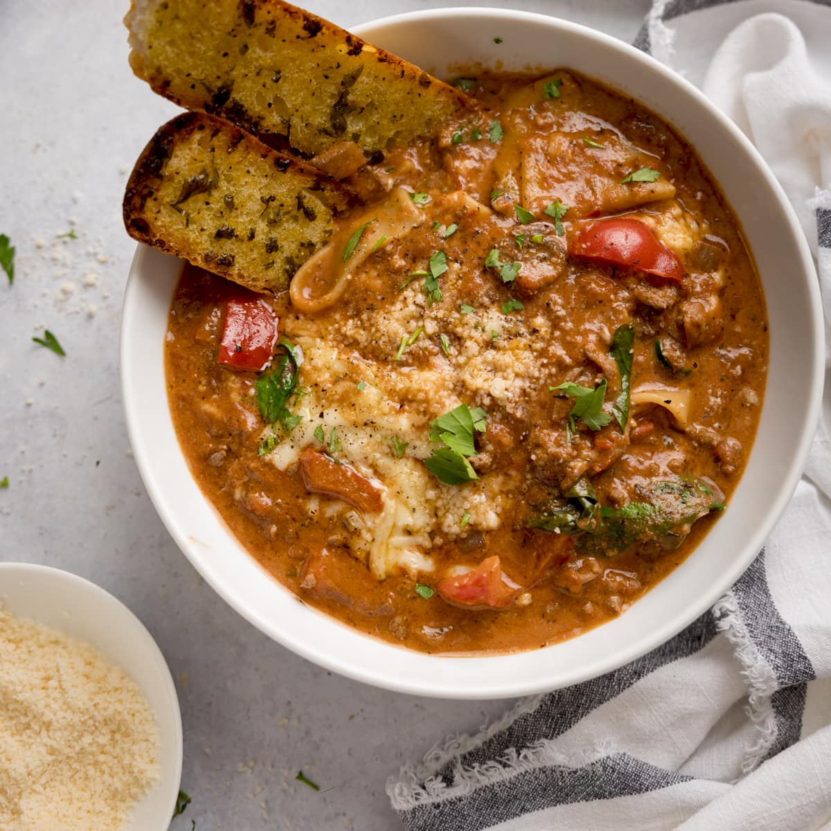 Square image of a white bowl filled with lasagne soup with pieces of garlic bread taken out. The bowl is on a light background next to a white napkin and a small bowl of grated parmesan.