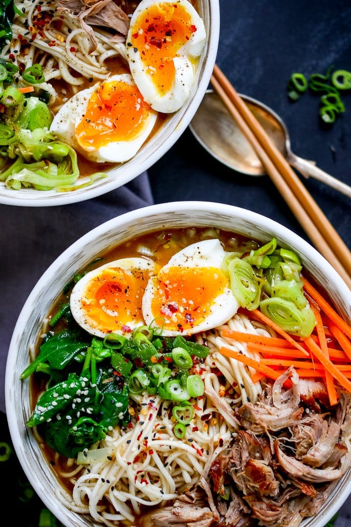 Overhead photo of 2 bowls full of pork ramen with pulled pork, noodles, spinach, runny eggs and broth on a blue background with a spoon and chopsticks