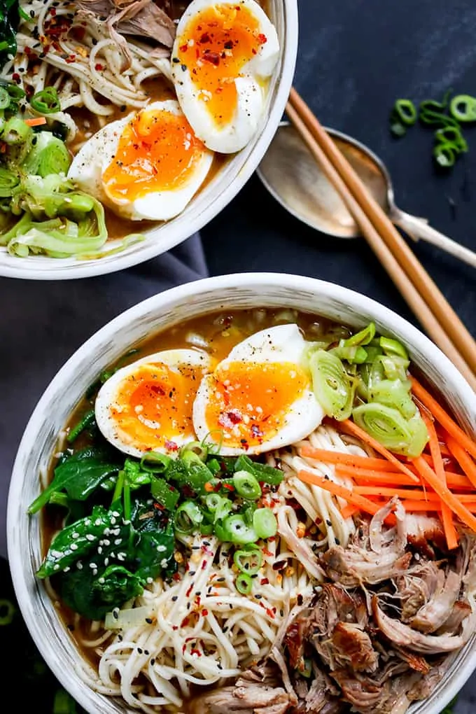 Overhead photo of 2 bowls full of pork ramen with pulled pork, noodles, spinach, runny eggs and broth on a blue background with a spoon and chopsticks