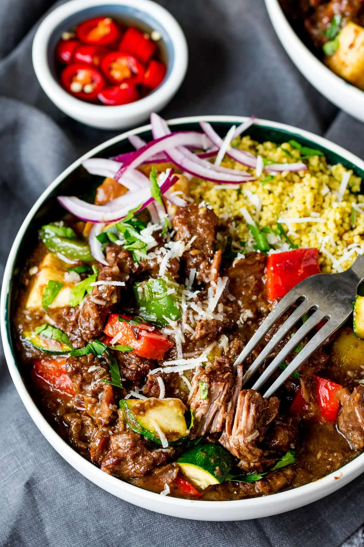 Slow cooked summer beef casserole in a bowl with peppers, courgette, red onion and couscous. Forkful being taken.