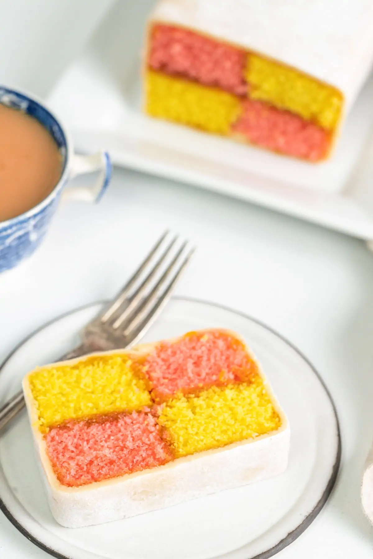 Slice of battenberg cake on a plate with a cup of tea and more battenberg in the background.