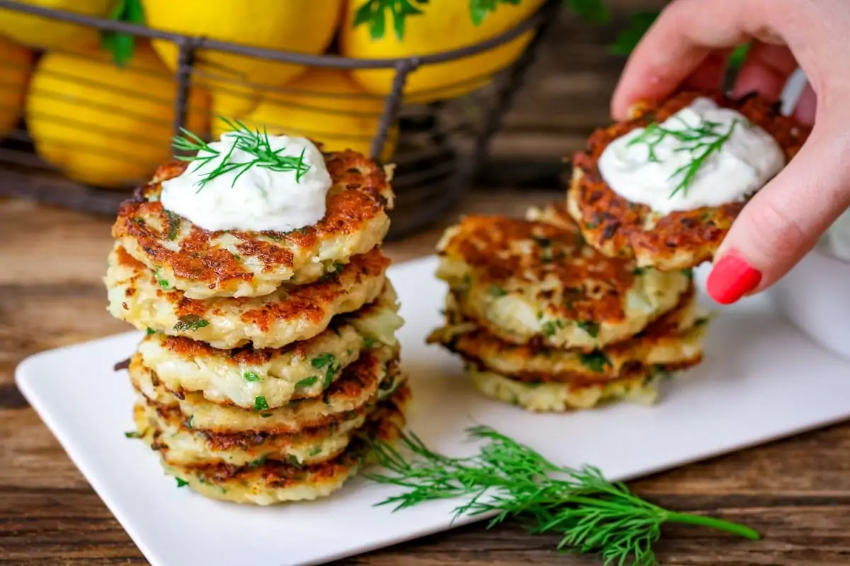 Two stacks of cauliflower fritters on a white plate. A hand is taking one of the fritters. In the background is a basket of lemons.