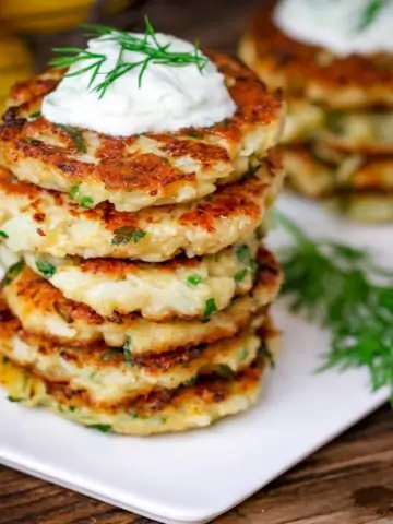 Square image of a stack of cauliflower fritters topped with tzatziki on a white plate on a wooden board.