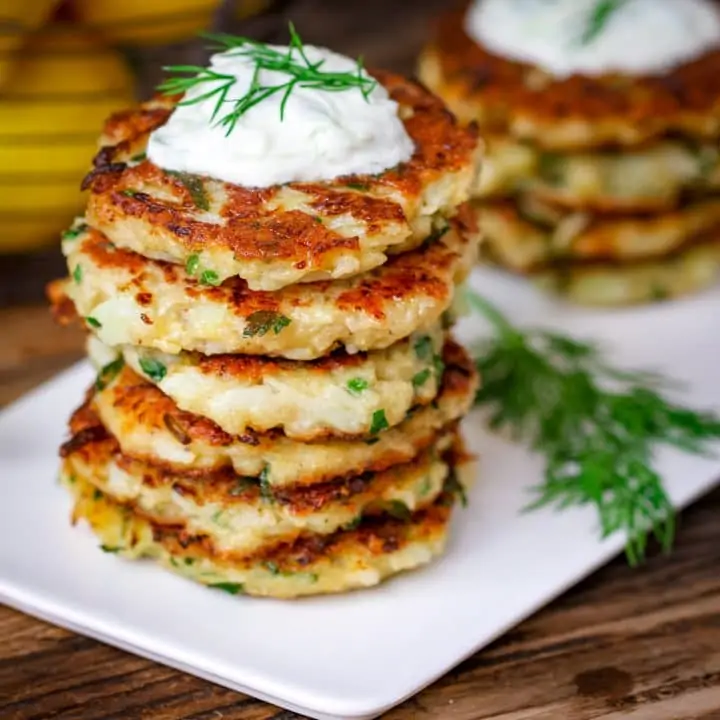 Square image of a stack of cauliflower fritters topped with tzatziki on a white plate on a wooden board.