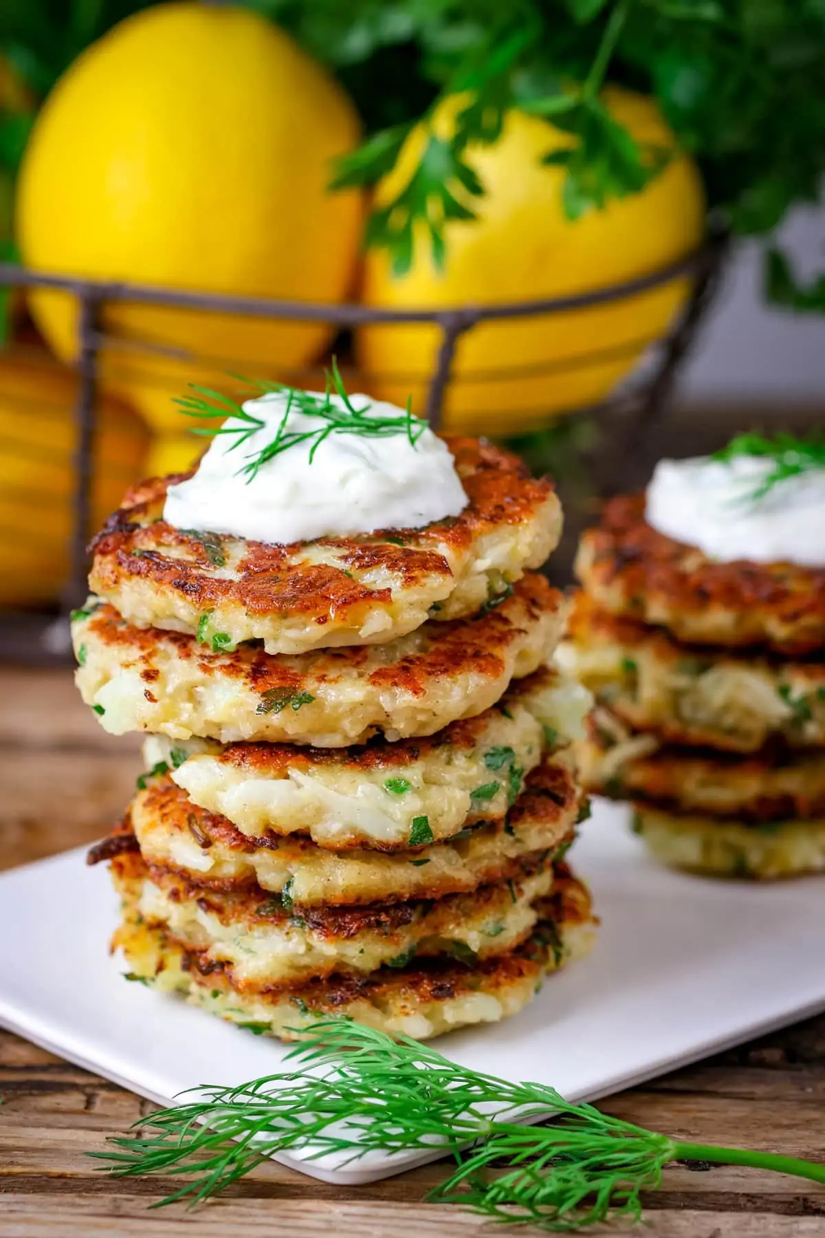A stack of cauliflower fritters on a white plate on a wooden board. More fritters plus a basket of lemons in the background.