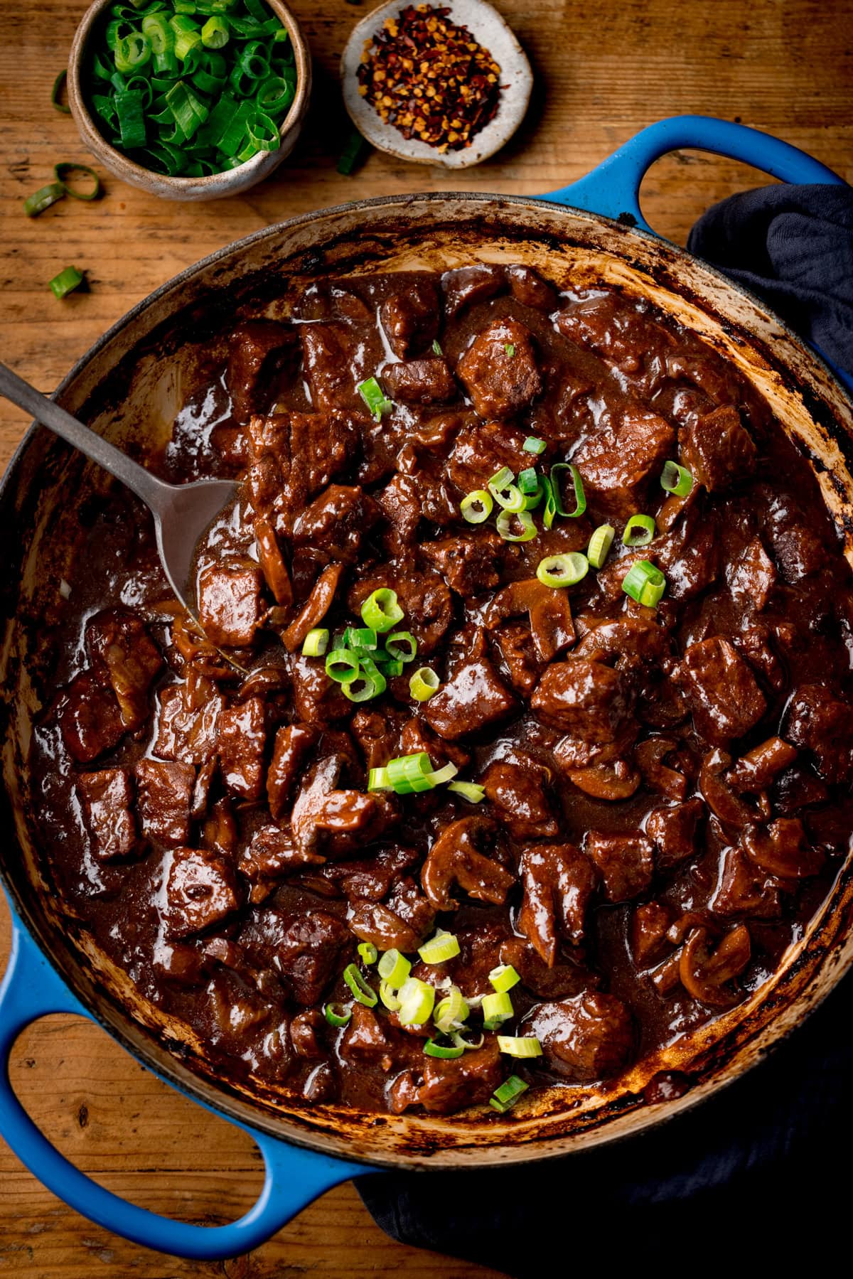Una cacerola de hierro fundido azul lleno de curry de carne china, cubierta con cebolletas en rodajas de primavera. Hay pequeños tazones de cebolletas picadas y copos de chile cerca. La sartén está en una superficie de madera.