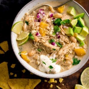 white chilli chicken con carne in a white bowl topped with lime wedges, avocado chunks and red onion. Tortilla crisps next to the bowl.