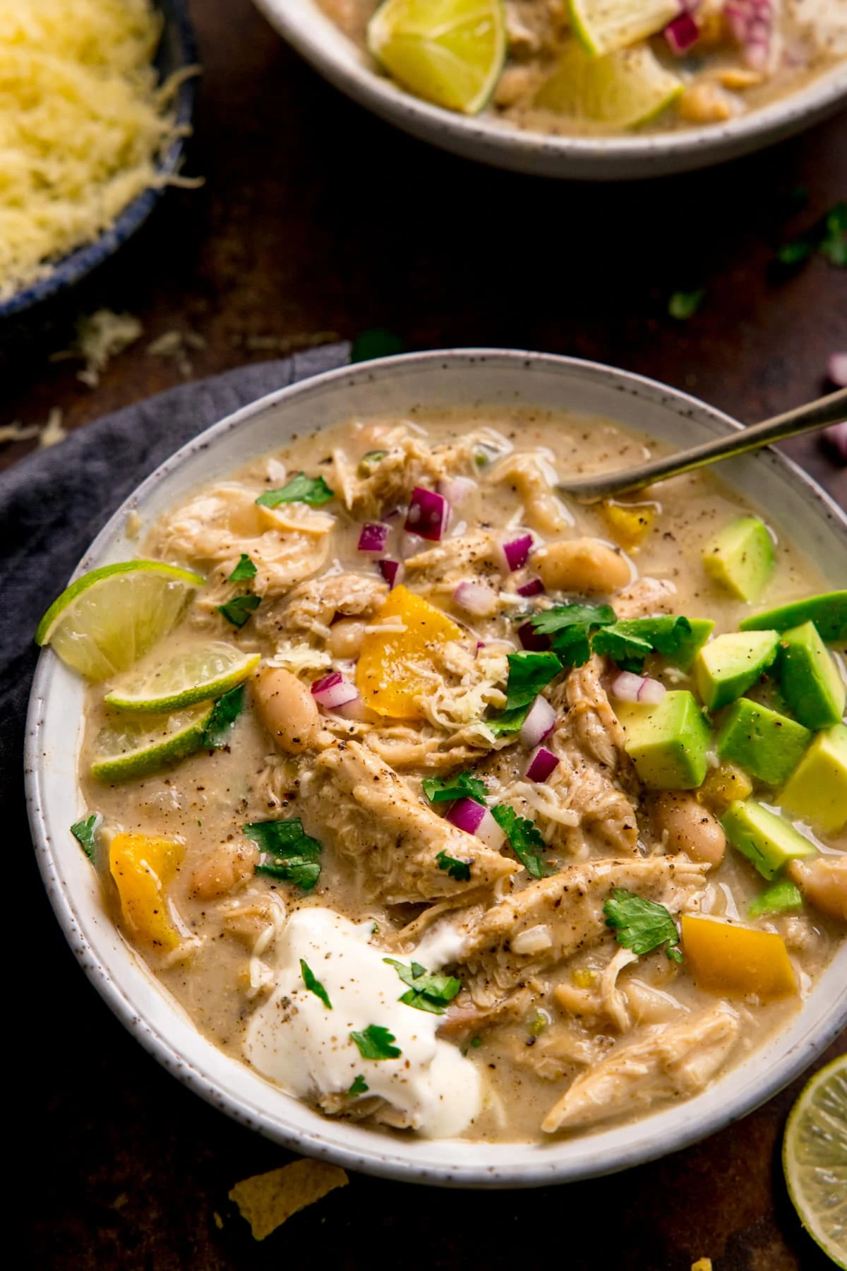 white chilli chicken con carne in a white bowl on a dark background. The white chilli chicken has toppings of avocado, red onion, sour cream, coriander and lime.