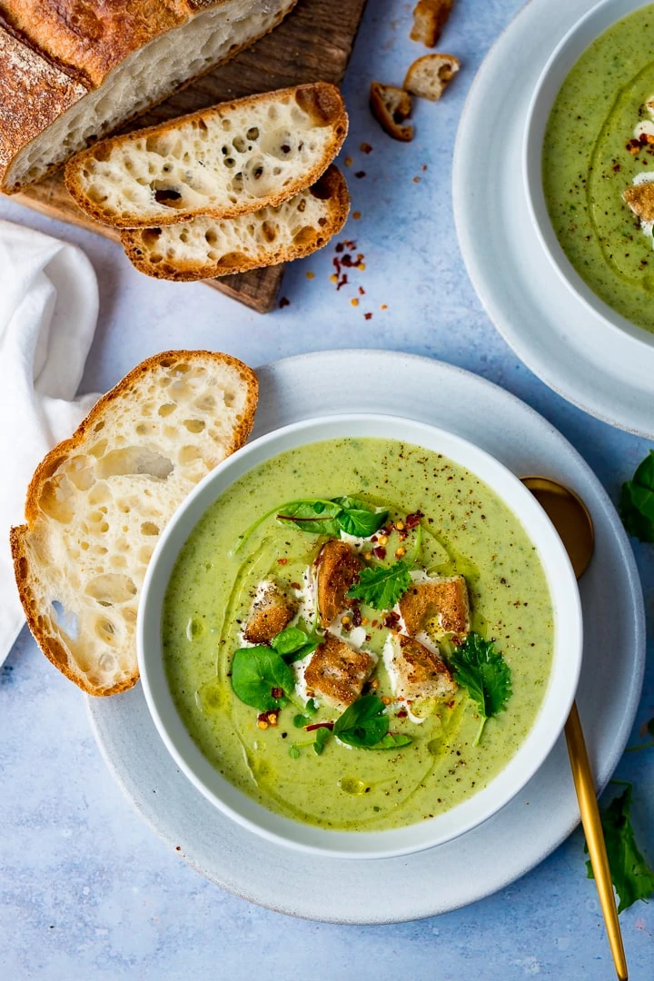 Broccoli soup in a bowl topped with croutons and baby salad leaves on a light blue background, served with freshly sliced bread