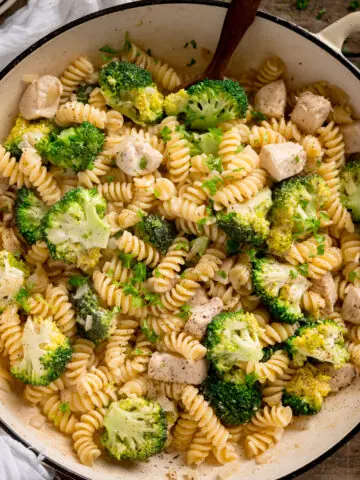 Overhead shot of chicken and broccoli pasta in a white cast iron pan with a wooden spoon sticking out. The pan is on a wooden table with a white napkin