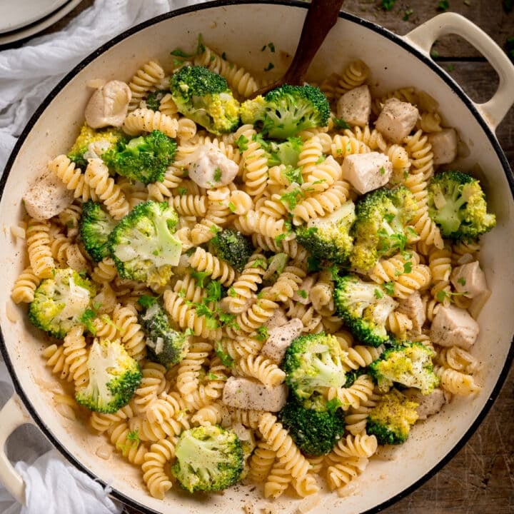 Overhead shot of chicken and broccoli pasta in a white cast iron pan with a wooden spoon sticking out. The pan is on a wooden table with a white napkin
