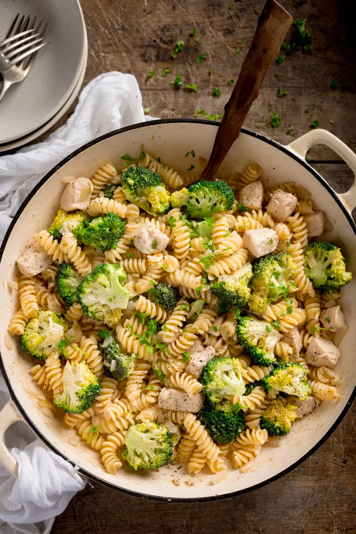 Overhead shot of chicken and broccoli pasta in a white cast iron pan with a wooden spoon sticking out. The pan is on a wooden table with a white napkin and bowls nearby.