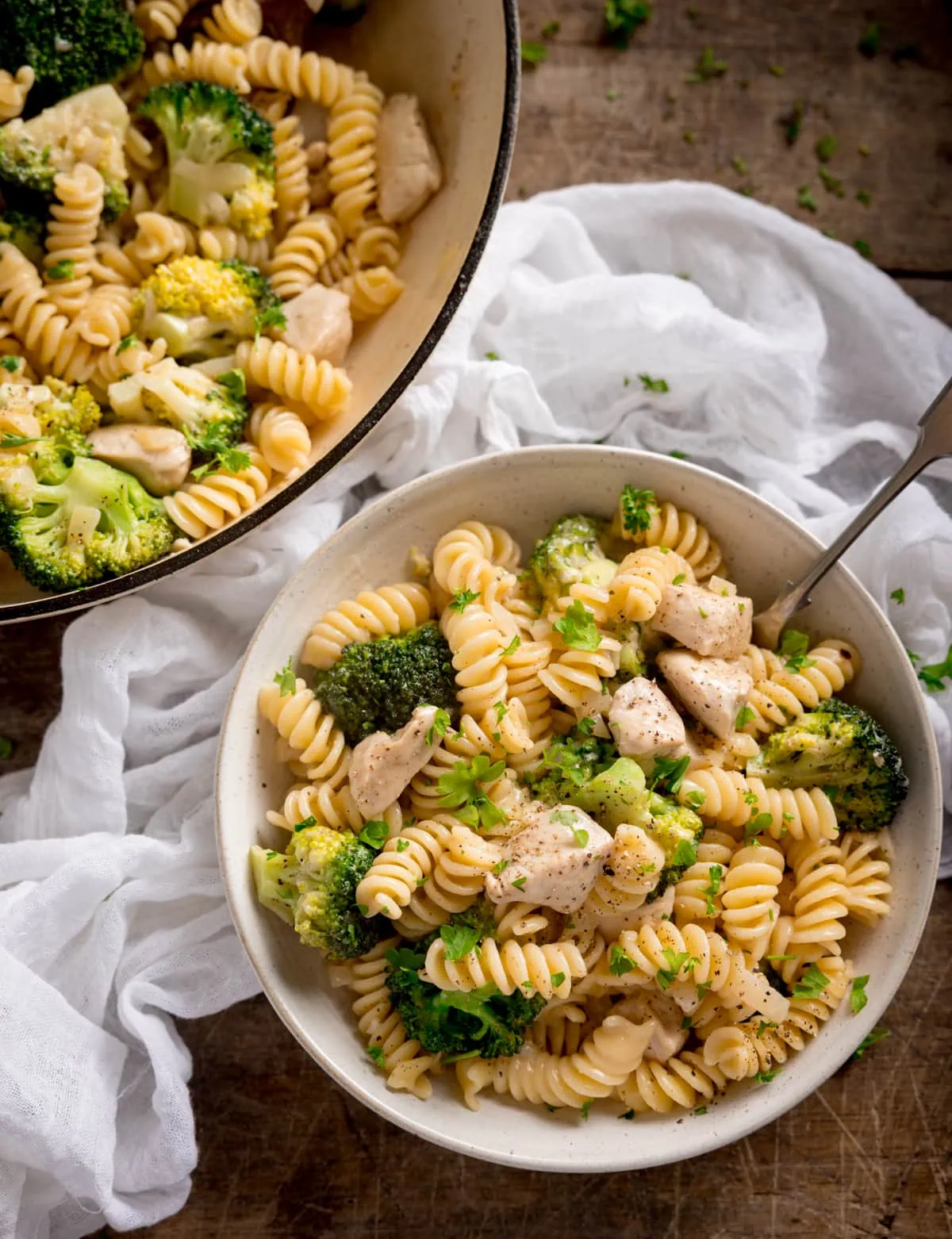 Overhead shot of chicken and broccoli pasta in a white bowl with a fork sticking out. The bowl is on a wooden table with the pan of pasta and a white napkin nearby.