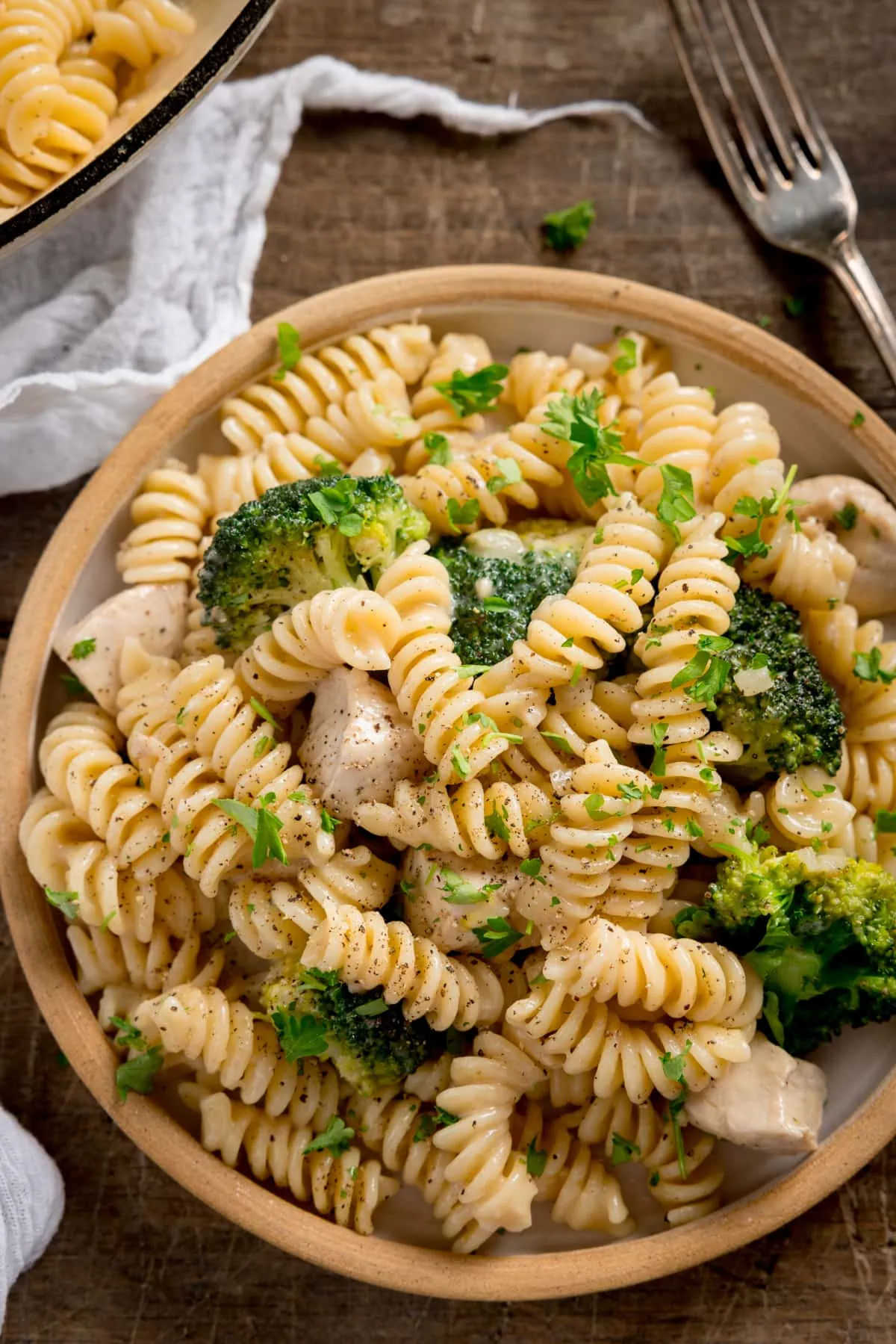 Overhead shot of chicken and broccoli pasta on a white plate with stone rim. The plate is on a wooden table with a white napkin and fork nearby.