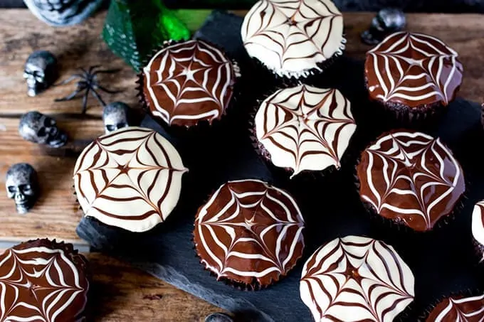 Chocolate Halloween cupcakes with spiderweb patterns in the topping placed on a dark slate background with toy skulls and spiders on a wooden board.