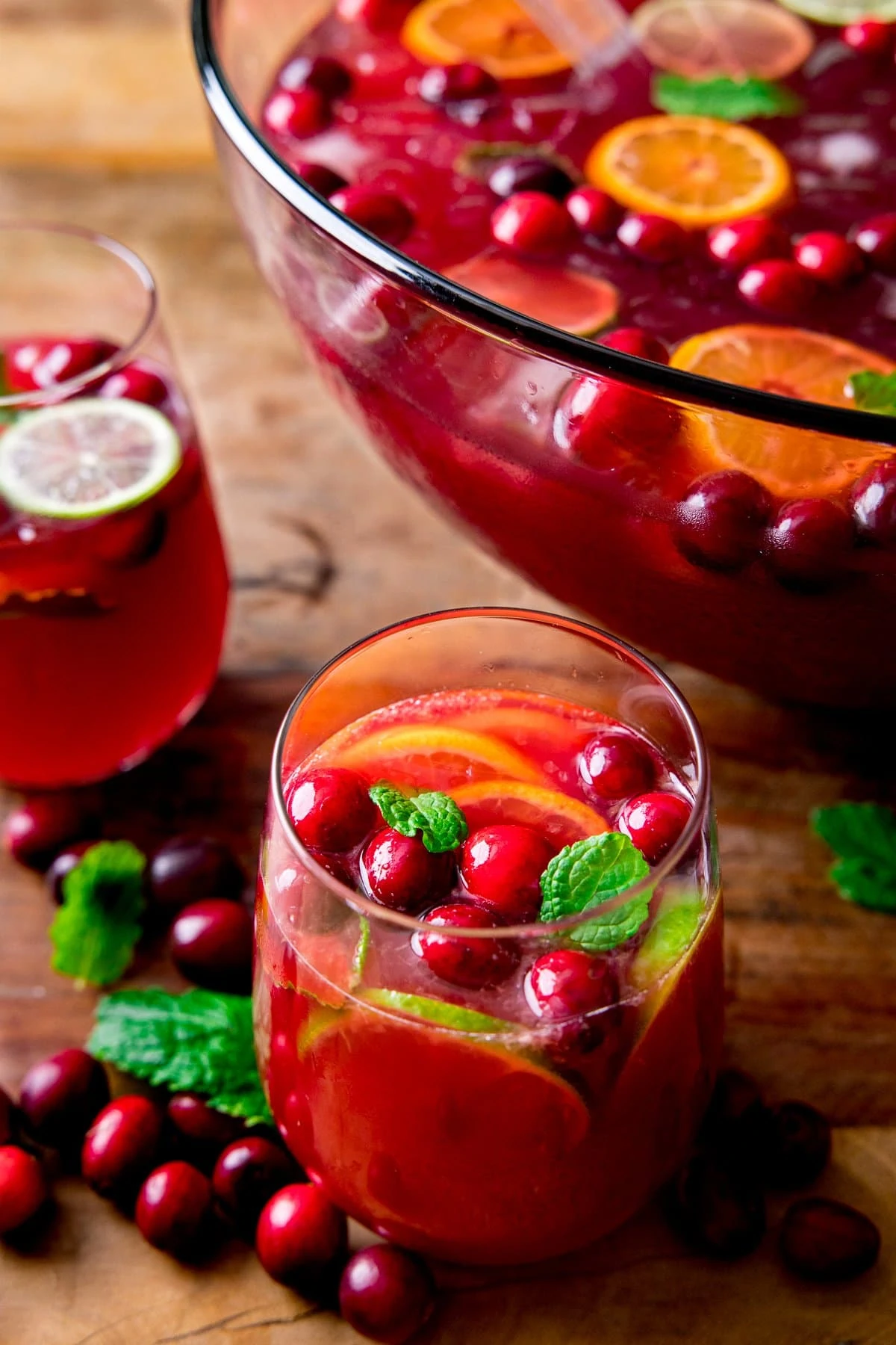 Glass of Christmas Punch in the foreground. Further one in the background, along with the bowl of punch. Punch is topped with citrus slices and cranberries.