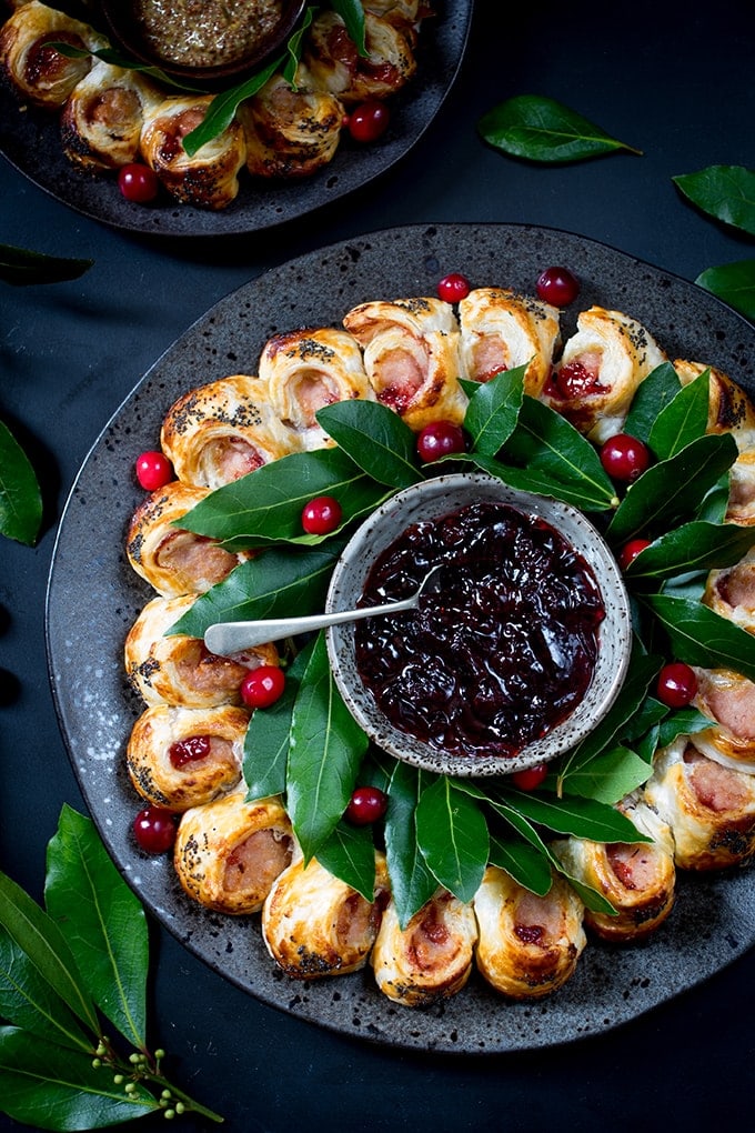 Festive sausage roll wreath topped with a honey mustard gaze and poppy seeds in a wreath shape with green leaves and a dish of cranberry sauce in the center