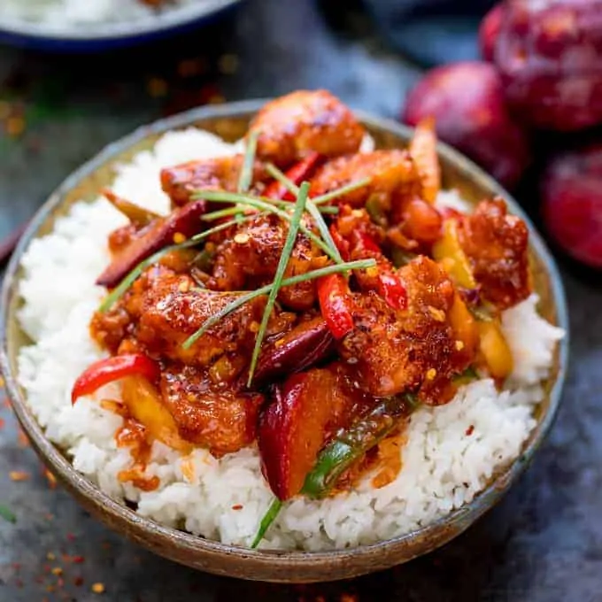 Chinese plum chicken stir fry and rice in a bowl. Whole plums and blue napkin next to bowl.