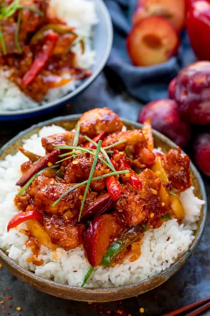 Chinese plum chicken stir fry and rice in a bowl. Further bowl at the top of the frame. Whole and halved plums and blue napkin next to bowls.