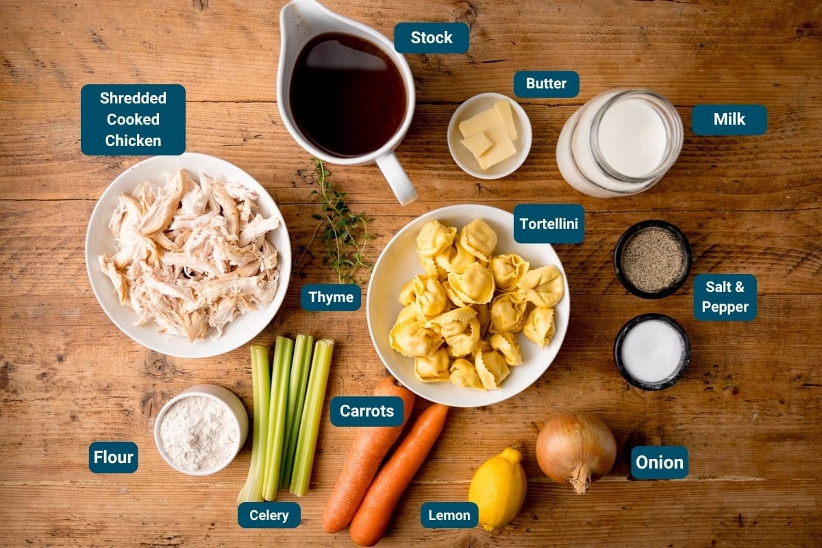 Ingredients for a Creamy Chicken and Tortellini Soup on a wooden table: shredded cooked chicken, stock, butter, milk, salt and pepper, onion, lemon, carrots, celery, flour, thyme and tortellini.