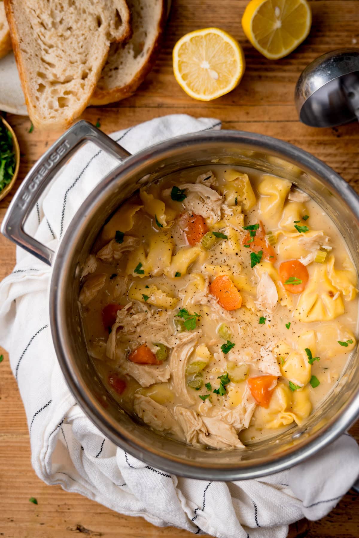 A pot of creamy chicken and tortellini soup with carrots and parsley sits on a wooden table, with slices of bread and lemon halves nearby.