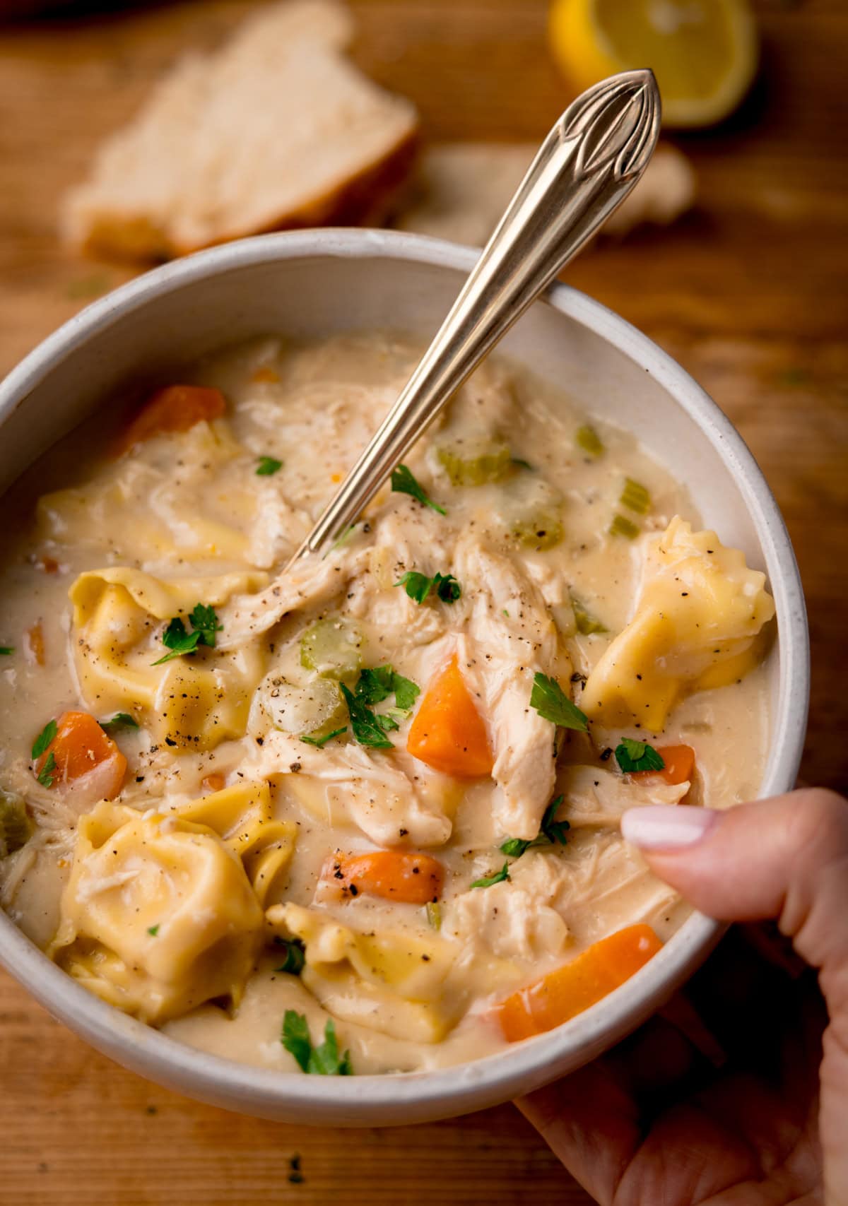 A bowl of creamy tortellini soup with chicken and vegetables, garnished with parsley, held in one hand with a spoon, bread, and a lemon slice in the background.