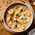 A bowl of creamy chicken and tortellini soup with carrots, celery, and parsley, served with slices of rustic bread on a plate.
