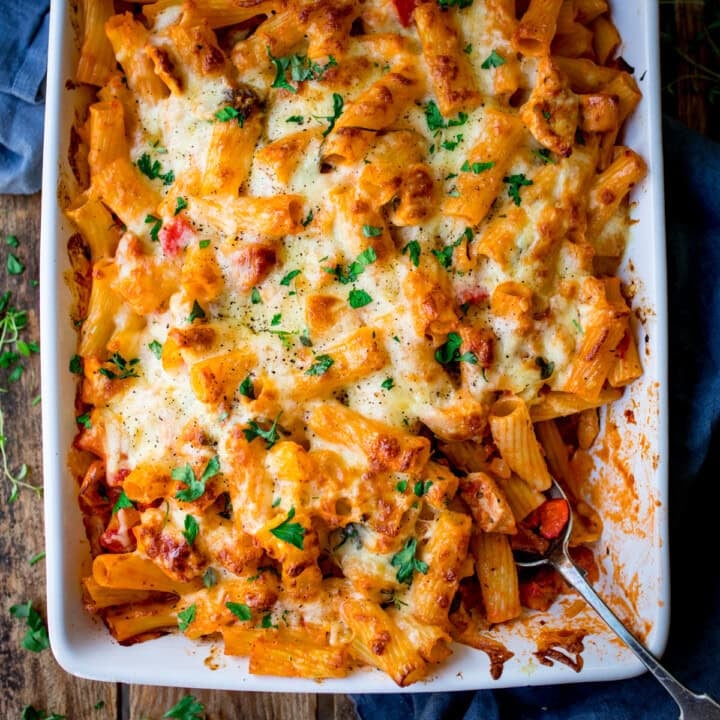 A cheesy chicken pasta bake served in a white dish, topped with melted cheese and fresh herbs. The pasta is mixed with tomato sauce and visible chunks of chicken and bacon, with a spoon on the side for easy serving.