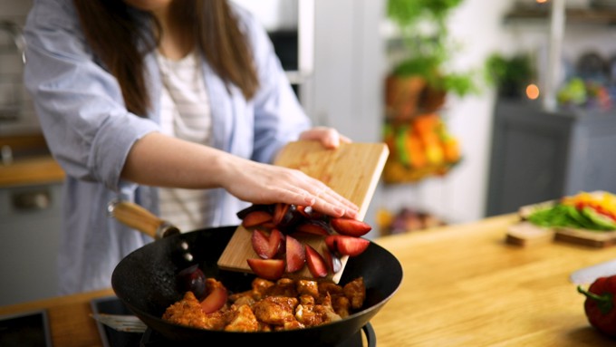 Plum being pushed from a chopping board into a wok of stir fried chicken by a lady in a blue shirt. Kitchen and fruit in background