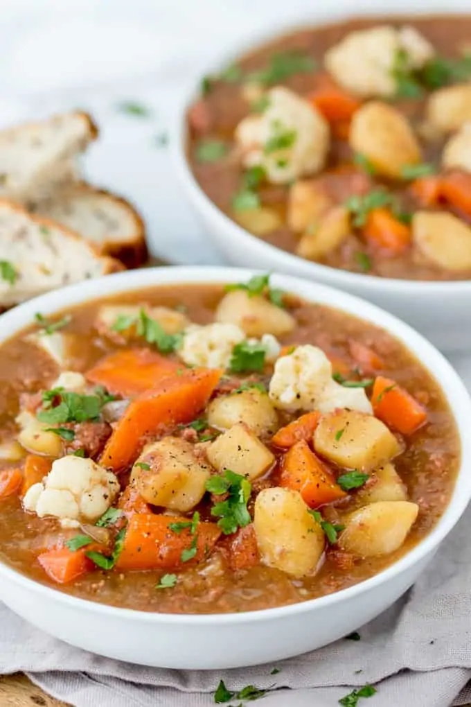 A picture of 2 bowls of corned beef hash soup on a table cloth with 3 pieces of bread in the background.