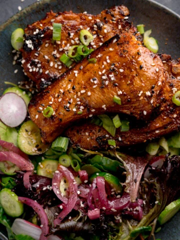 Overhead square image of 3 marinated and cooked lamb cutlets on a dark blue plate next to a side salad. The lamb cutlets are sprinkled with sesame seeds and chopped spring onions.