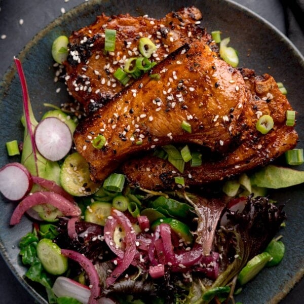 Overhead square image of 3 marinated and cooked lamb cutlets on a dark blue plate next to a side salad. The lamb cutlets are sprinkled with sesame seeds and chopped spring onions.