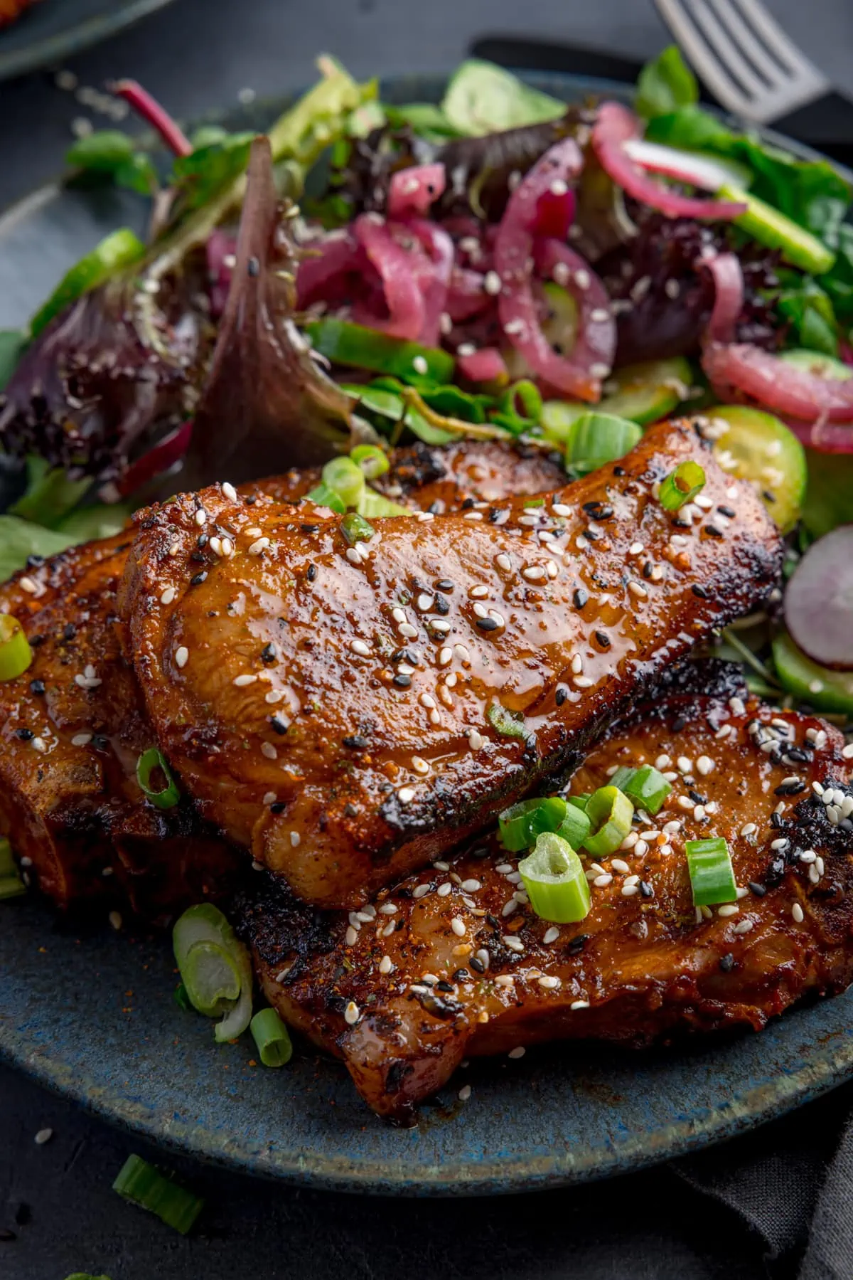 3 marinated and cooked lamb cutlets on a dark blue plate next to a side salad. The lamb cutlets are sprinkled with sesame seeds and chopped spring onions.