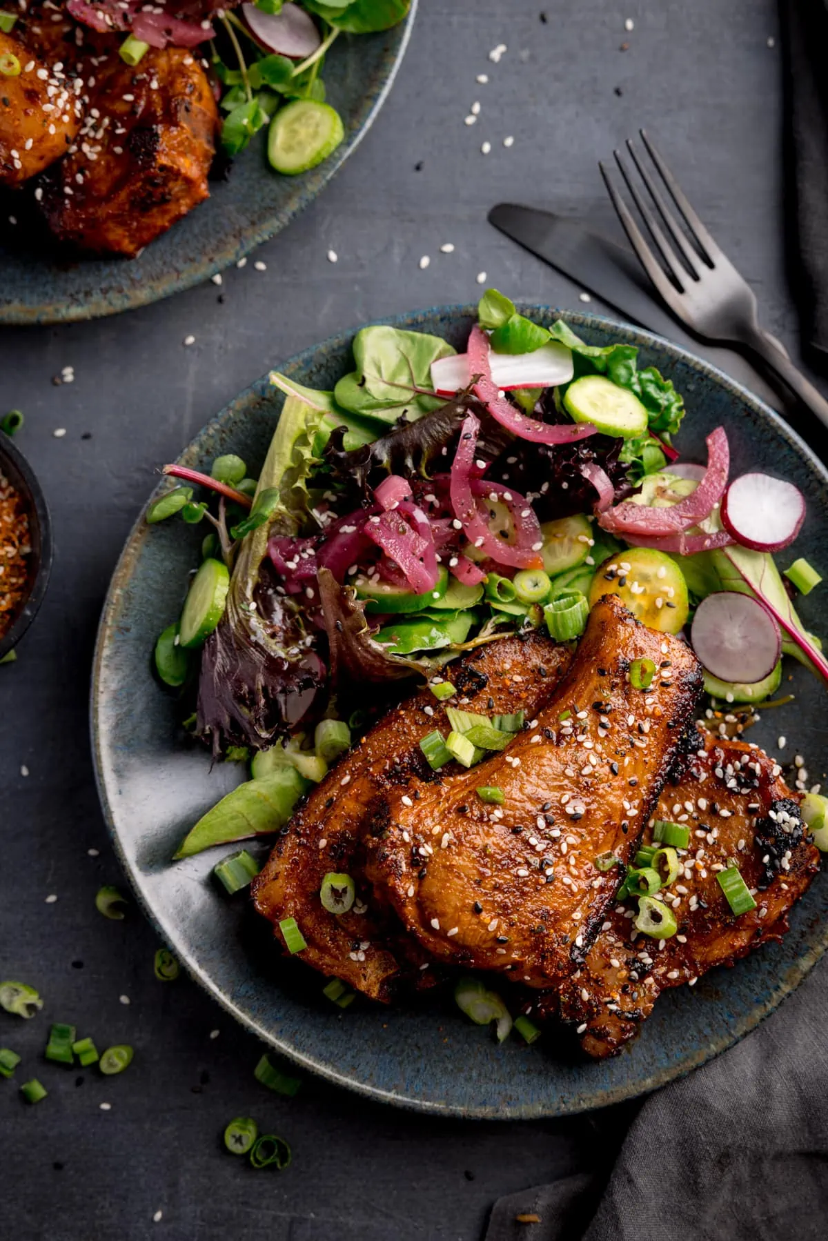 Overhead image of 3 marinated and cooked lamb cutlets on a dark blue plate next to a side salad. The lamb cutlets are sprinkled with sesame seeds and chopped spring onions. The plate is on a dark grey background, next to a dark knife and fork and a further plate of lamb cutlets.