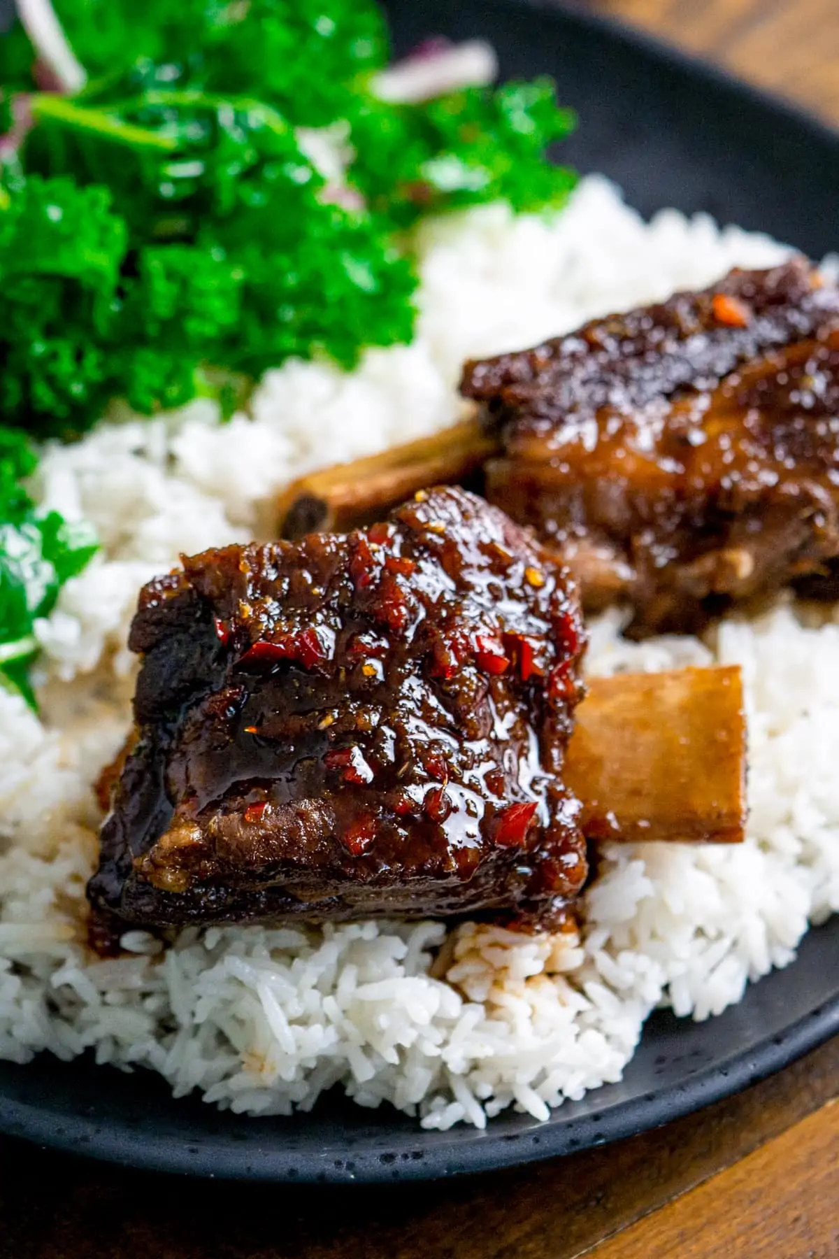 Slow cooked short ribs with chilli glaze on a bed of rice on a dark plate. Kale is also on the plate in the background.