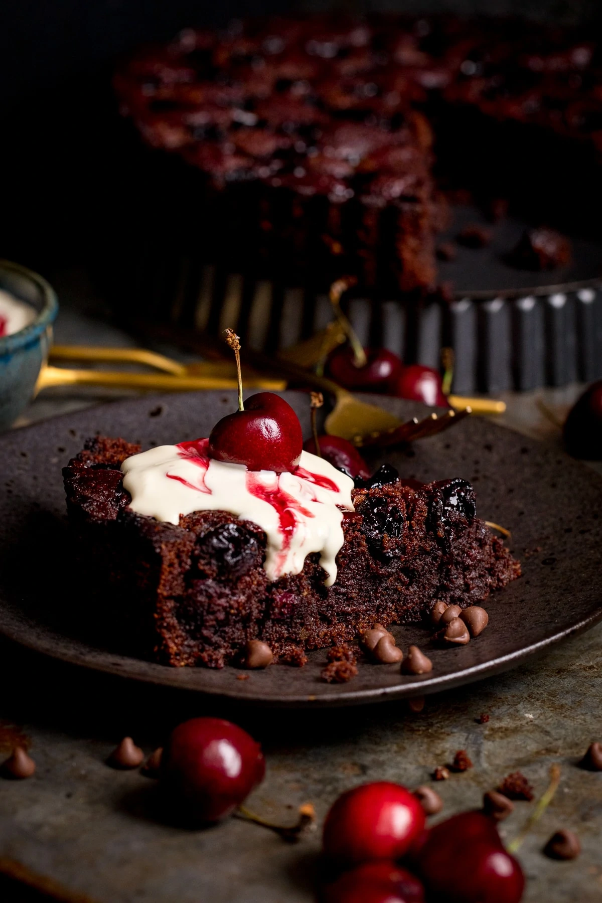 Chocolate Cherry Cake slice on a dark plate. Cherries in foreground, rest of cake in background.