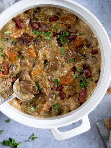 Overhead image of creamy pork and bacon casserole in a white casserole pan, on a light blue surface. There is a spoonful of the casserole being taken. There is some torn bread and parsley on the table, around the casserole pan.