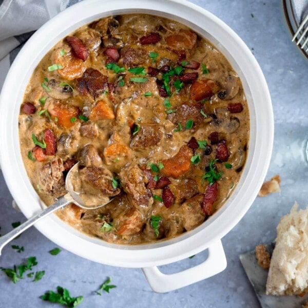 Overhead image of creamy pork and bacon casserole in a white casserole pan, on a light blue surface. There is a spoonful of the casserole being taken. There is some torn bread and parsley on the table, around the casserole pan.