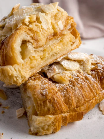 An almond croissant sliced in half and stacked on a white plate on a white background. There is a pale pink napkin in the background.