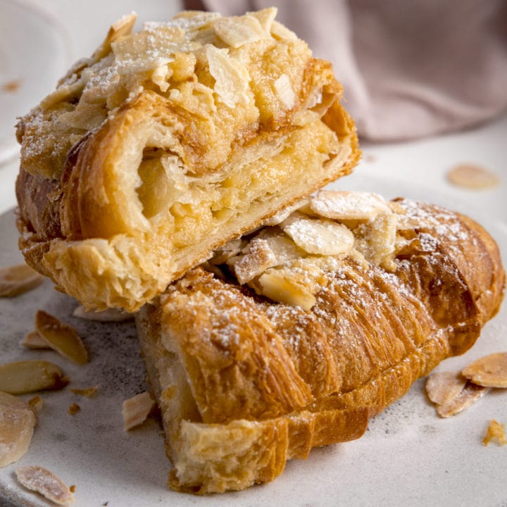 An almond croissant sliced in half and stacked on a white plate on a white background. There is a pale pink napkin in the background.