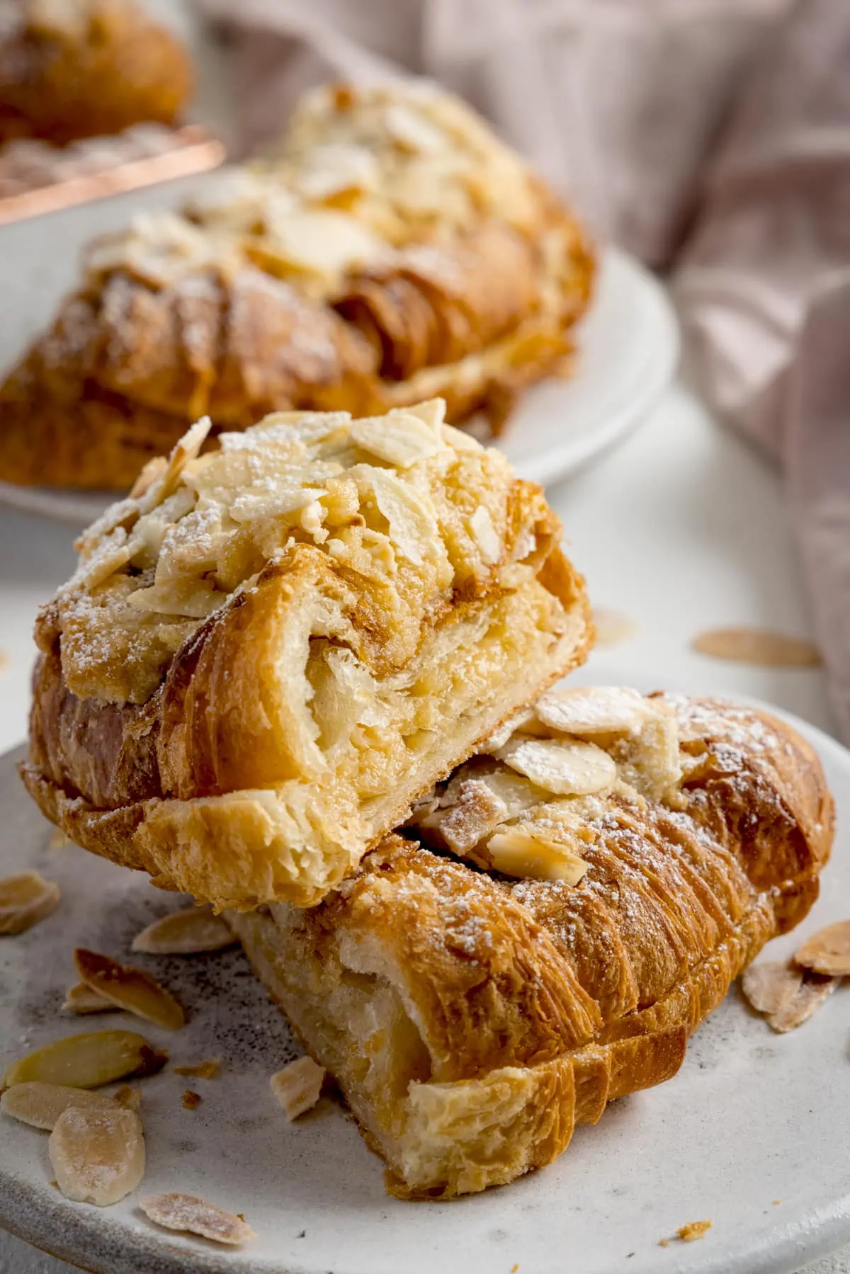 An almond croissant sliced in half and stacked on a white plate on a white background. There are further croissants and a pale pink napkin in the background.