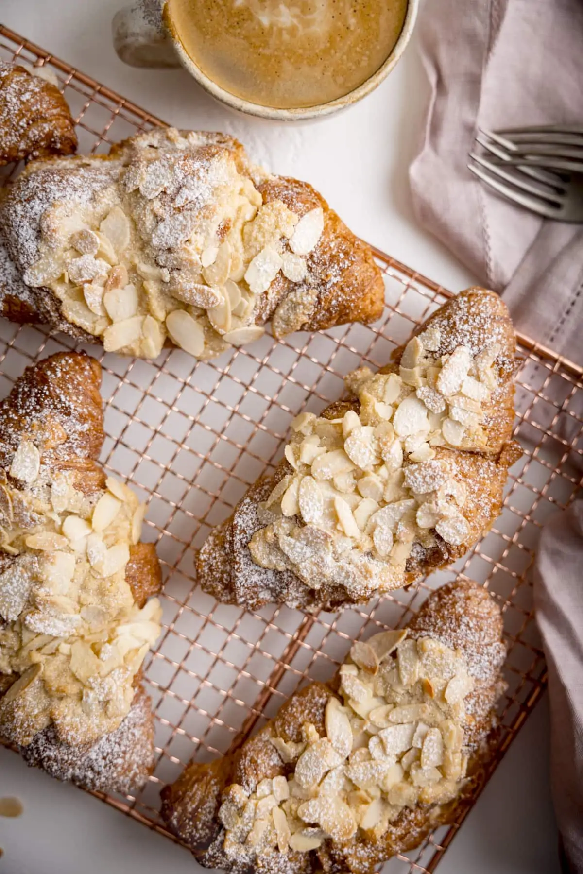 Overhead image of 4 almond croissants on a gold wire cooling rack, on a white background. There is. cup of coffee and a dusty pink napkin at the top of the frame.