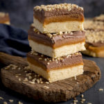 3 pieces of salted caramel millionaire's shortbread piled on a rustic wooden board, against a dark background.