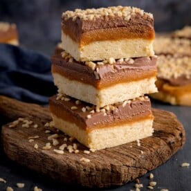 3 pieces of salted caramel millionaire's shortbread piled on a rustic wooden board, against a dark background.