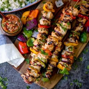 Square overhead shot of Mexican chicken kebabs piled on a wooden board. Salsa and rice in the background.